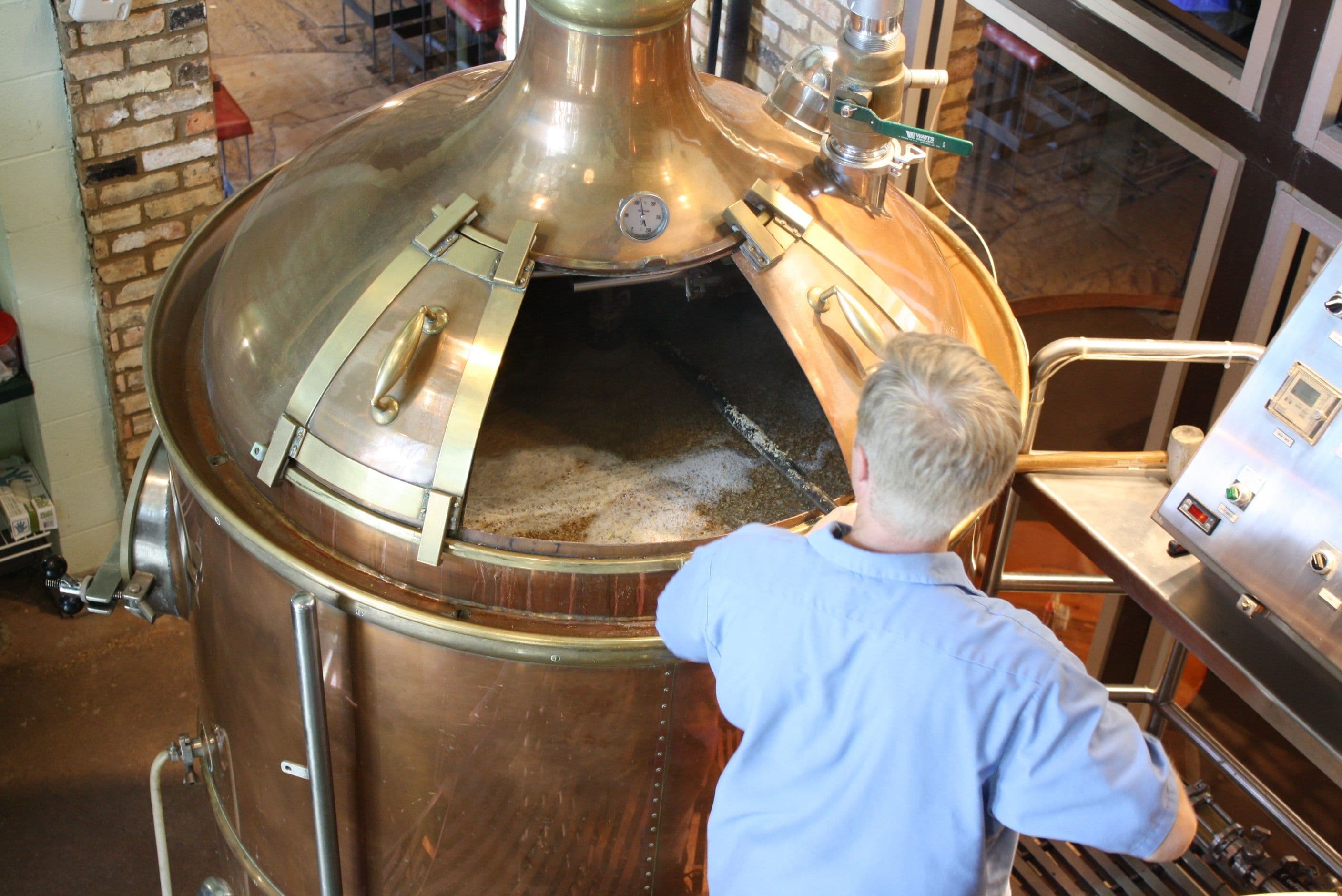 A person inspects boiling liquid in a large, shiny copper brewing vat.