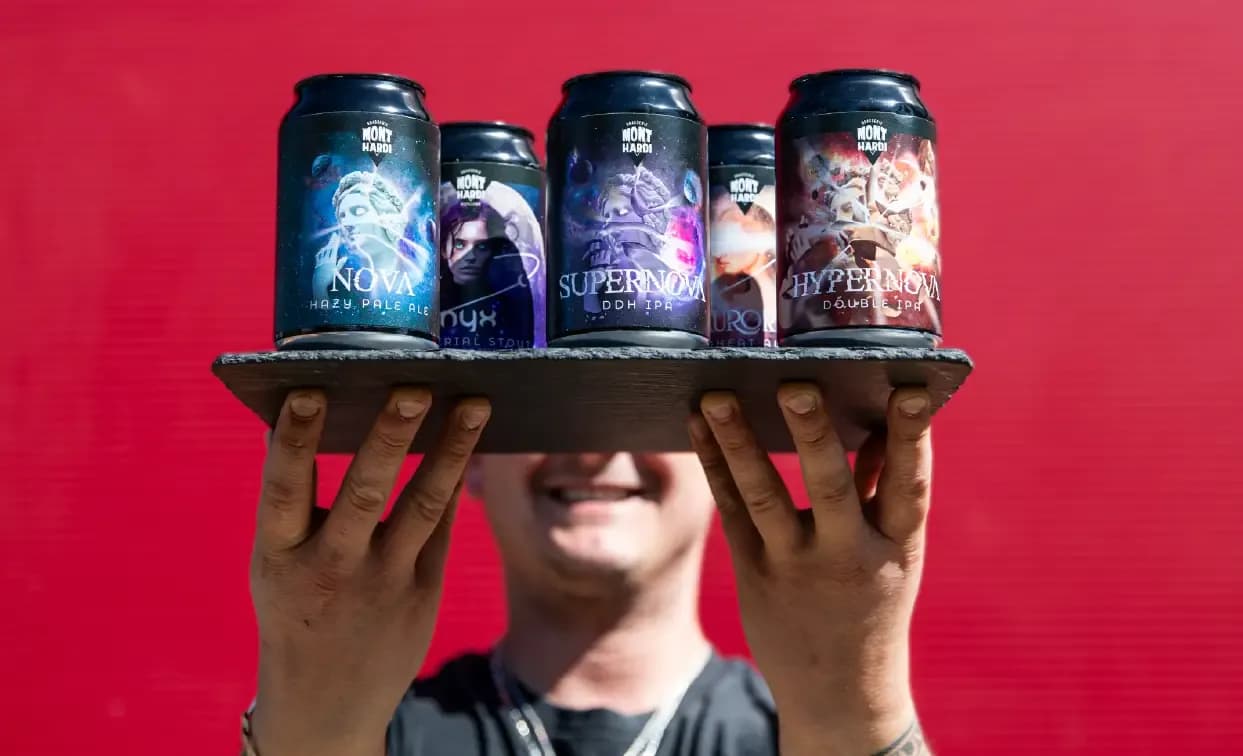 Smiling person holds a tray displaying five dark-labeled beverage cans against a red background.