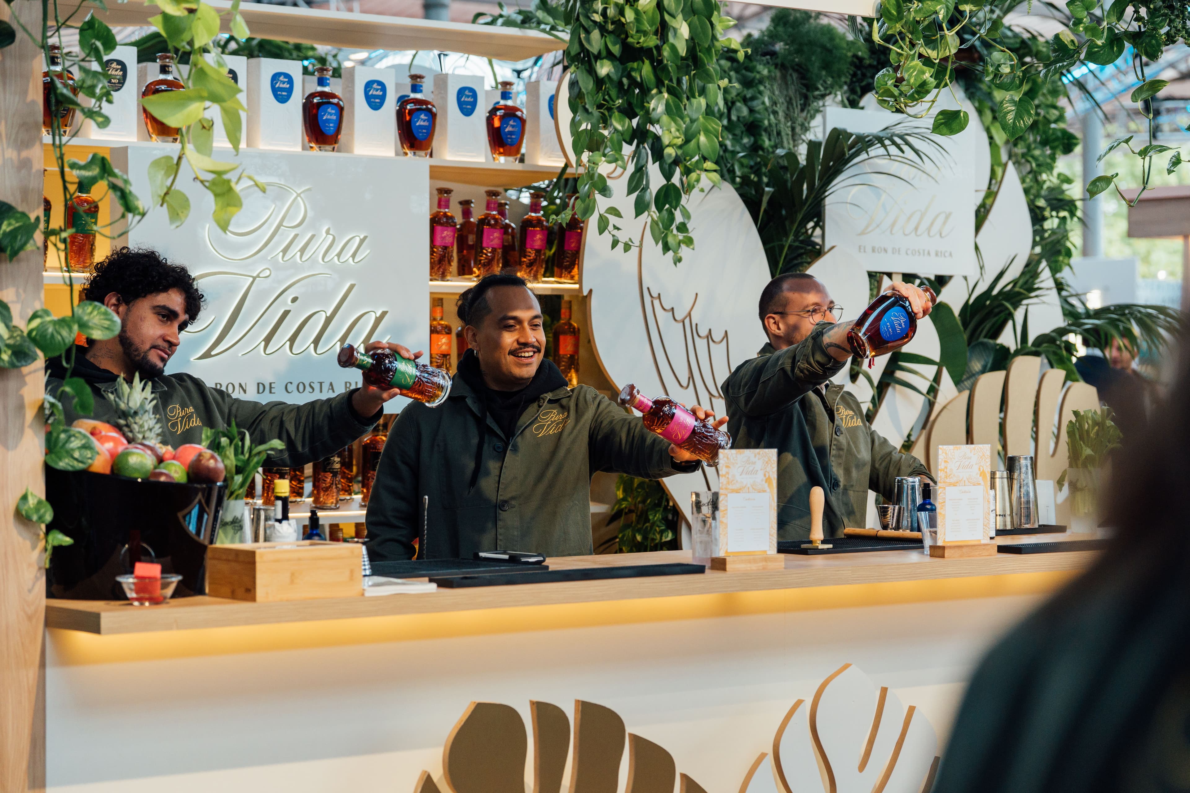 Three bartenders cheerfully shake colorful cocktails behind a lively bar adorned with plants and a 'Pura Vida' sign.