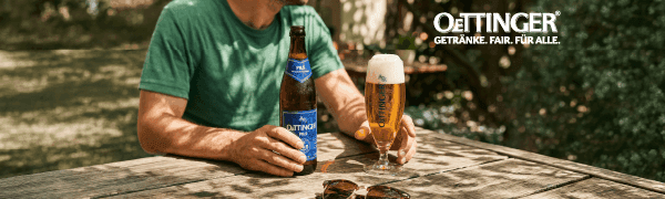 Man in green shirt holds Oettinger beer bottle and glass on stone table outdoors.