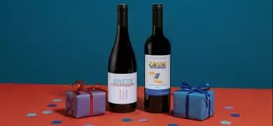 Two wine bottles and two wrapped gifts sit on a red surface with scattered confetti.