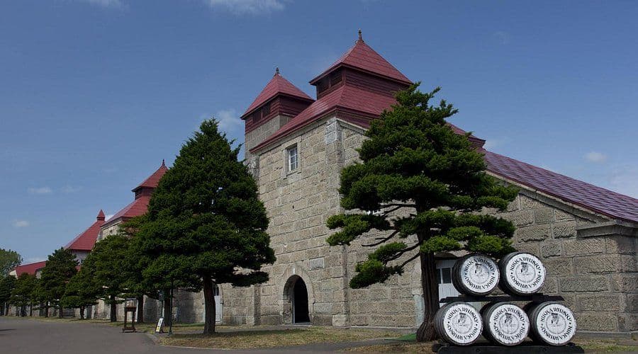 Historic stone building with red roofs, green trees, and stacked barrels in front.