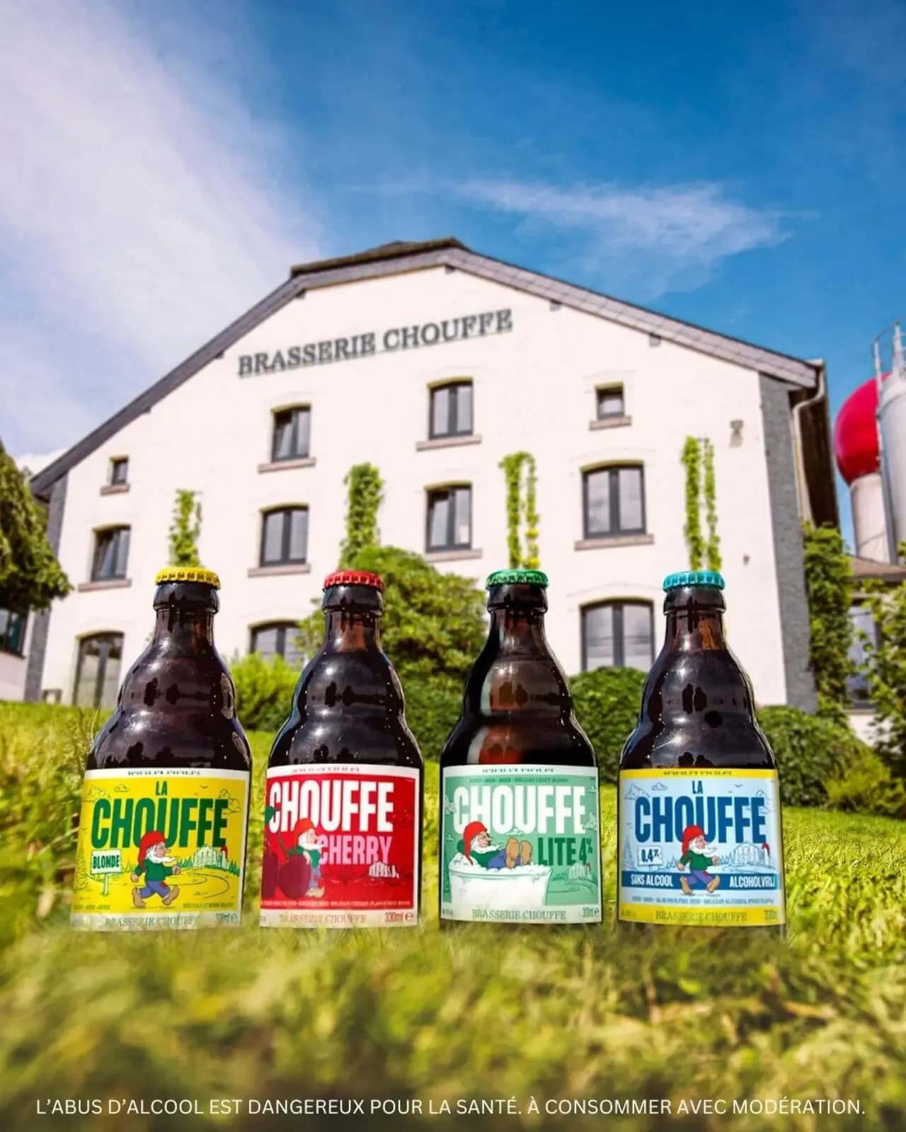Four distinct Chouffe beer bottles stand in grass before the Brasserie Chouffe building.