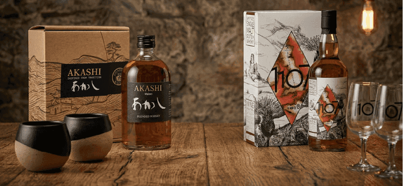 Two Japanese whiskey bottle sets, each with packaging and distinctive glasses, on a wooden table.