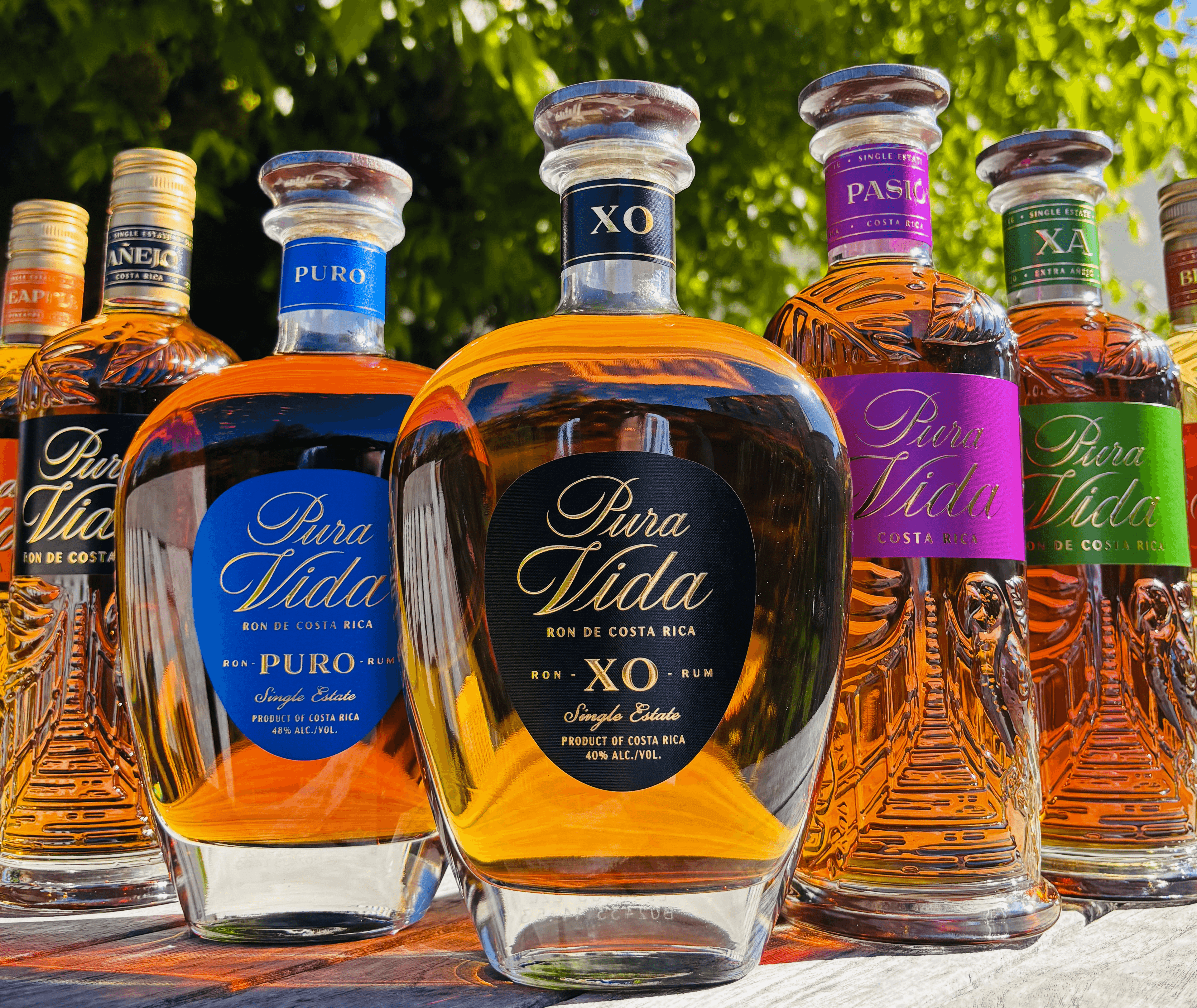 Several Pura Vida XO liquor bottles with amber liquid and colorful labels arranged outdoors.