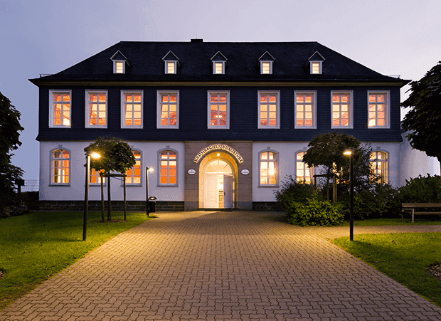Historic building with warmly lit windows at dusk, paved path leads to entrance.