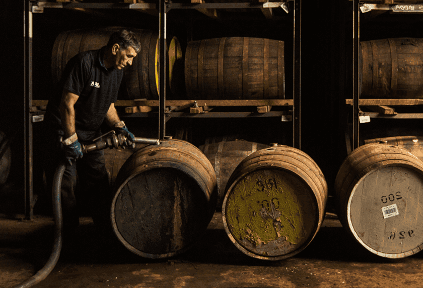 Man siphoning liquid into oak whiskey barrels in a dimly lit aging cellar.