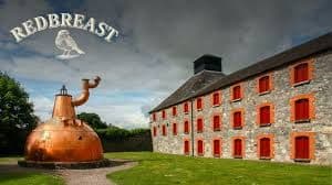 Large copper pot still and historic stone building at a Redbreast whiskey distillery.