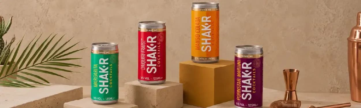 Four SHAK-R beverage cans in vibrant colors, displayed with a palm leaf and bar tools.