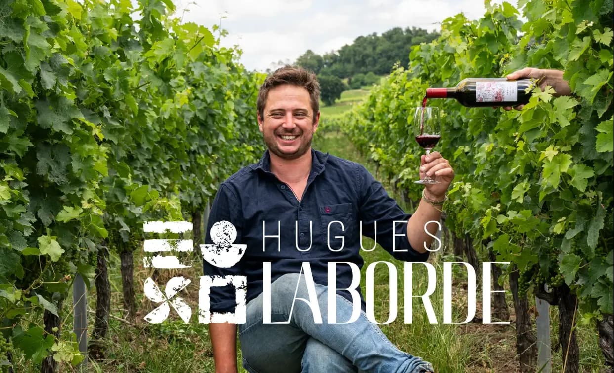 Hugues Laborde smiles in a vineyard as red wine is poured into his glass.