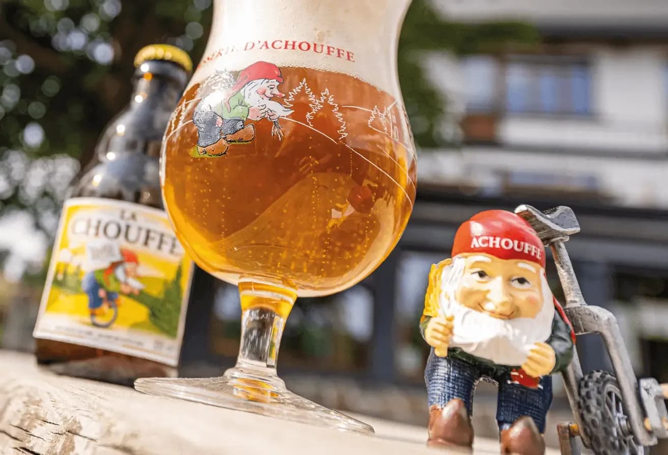 La Chouffe beer glass, bottle, and gnome figurine with bicycle outdoors.