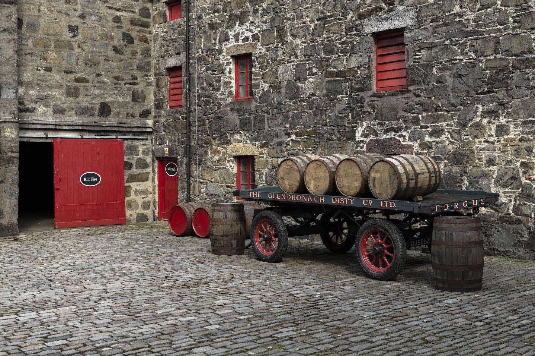 A rustic wooden cart with barrels on cobblestones before an old stone distillery building.