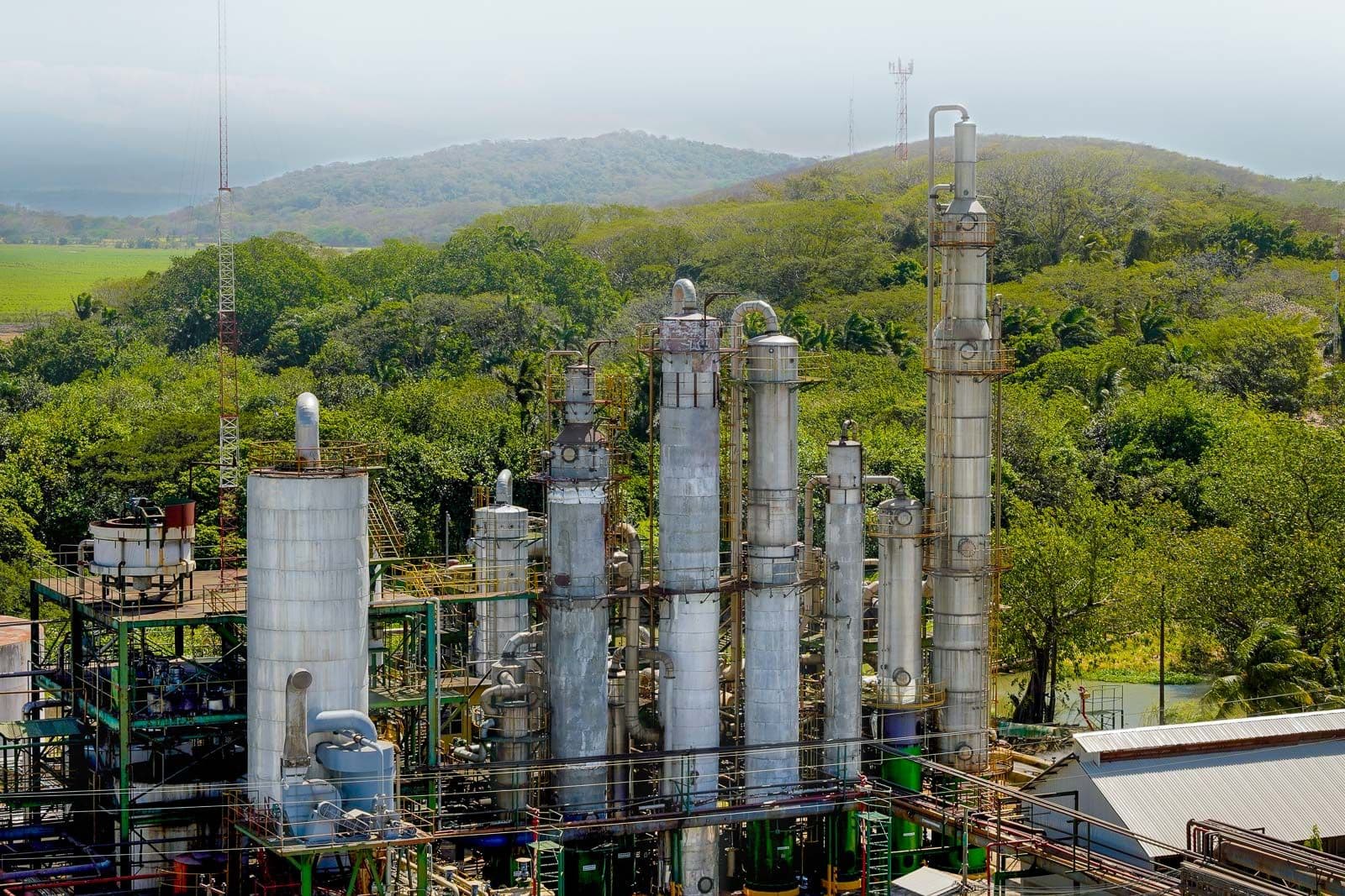Industrial chemical plant with tall distillation towers nestled among green hills under a hazy sky.