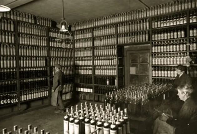 Workers in a vintage bottling plant surrounded by shelves of filled bottles.