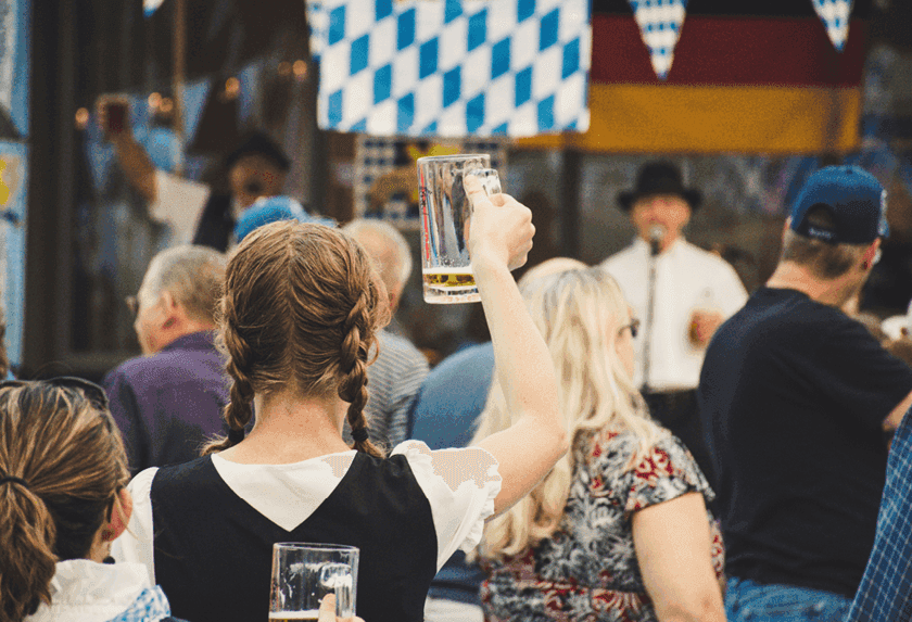 Woman in dirndl raises beer glass at a lively German festival with flags.