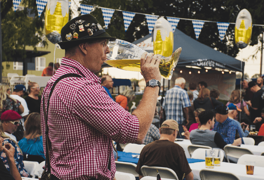 Man in traditional German hat and suspenders drinks from a large beer boot at an outdoor festival.